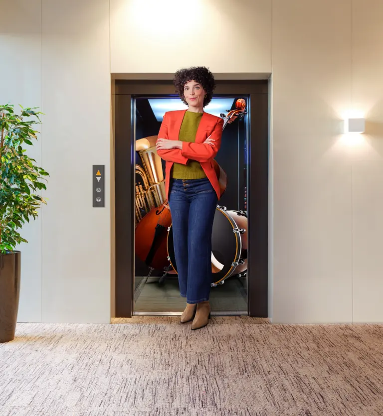 Mulher de braços cruzados à porta de um elevador aberto, com instrumentos musicais no interior, num corredor moderno com planta decorativa ao lado.